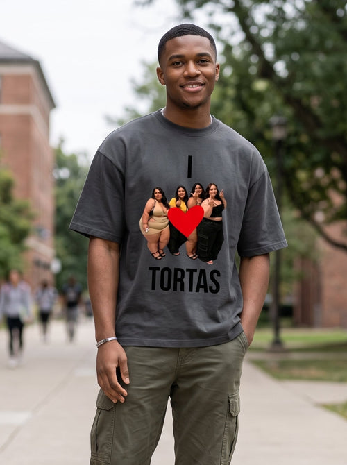 A man standing on a campus walkway wearing a dark gray graphic t-shirt with a 'I [photo of people] [red heart] TORTAS' design, olive green cargo pants, and dark sneakers.
